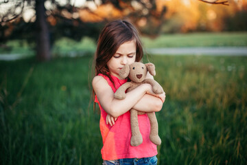 Sad upset Caucasian girl hugging toy. Child embrace soft plush bear in park outdoor. Lost lonely...