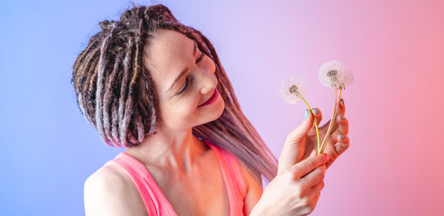 A young happy woman with dreadlocks is holding a white dandelion on a blue and pink background. Concept of a fun summer mood