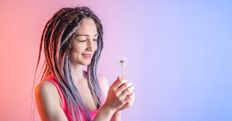 Obraz premium A young happy woman with dreadlocks is holding a white dandelion on a blue and pink background. Concept of a fun summer mood