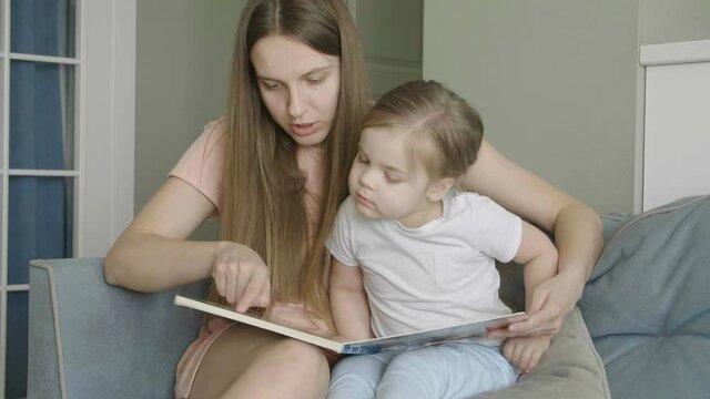 Caring single parent young mother babysitter hug happy cute little preschool child girl kid daughter hold learn read book fairy tale story bonding enjoying family lifestyle hobby at home sit on couch