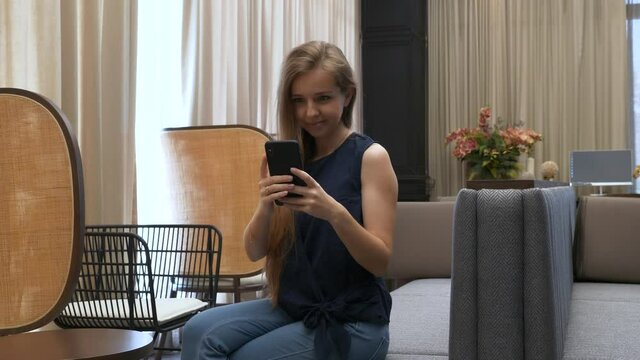 Happy Beautiful Young Girl Sitting on the Couch and Using the Mobile Phone, Smiling, Texting, Sharing Messages on Social Media