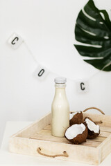 Cracked coconut and bottle of coconut milk on rustic wooden tray. Wooden Tray With Coconut and milk. Palm leaf and letters garland on the white wall. Selective focus.