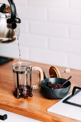 Loose hibiscus tea brewing in french press with hot water. Pouring water from metallic teapot. Bright kitchen interior. Wooden complete kitchen with gas oven