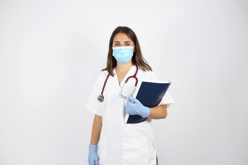 Young beautiful woman standing over white isolated background wearing her doctor uniform and smiling with her mask and notebook