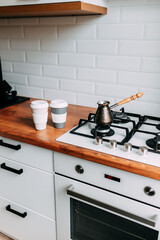Coffee brewing at home in jezve on gas oven. Coffee cups to go on the wooden table. White kitchen interior. Wooden complete kitchen with gas oven