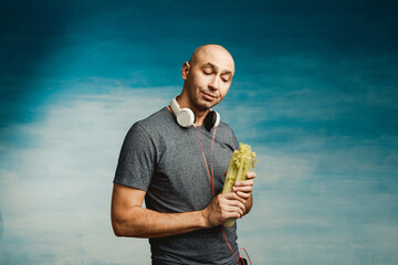 Funny bald man in headphones on his neck lustfully looks at the celery in his hands (on a blue...