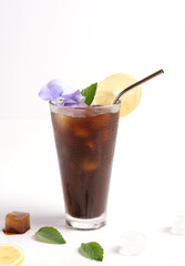 A glass of ice coffee stands on a white background. Coffee is decorated with an edible flower, a leaf of mint and a slice of lemon. Nearby are ice cubes and a slice of lemon. White background.