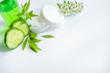 Face cream in a glass jar, slices of fresh cucumbers, green refreshing gel in a bottle on a white background. Fresh leaves and white flowers. Spa treatments. View from above. Copy space