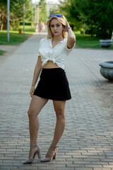 Obraz premium A young blonde in a black skirt and white blouse. Long beautiful legs. Standing on the sidewalk in the Park. Wearing sunglasses.