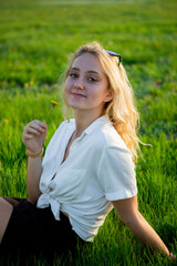 A young blonde is sitting in a clearing and holding a dandelion. Green grass. A girl poses in a meadow.