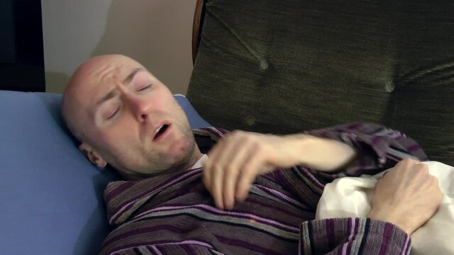 A man lay sick at home wearing a dressing gown and resting on his couch. He coughs and wipes his nose. Staying at home and self-isolating so that he does not spread the virus to other people.