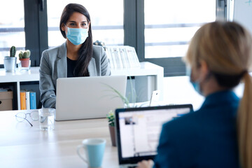 Two business women wearing a hygienic face mask while work with laptops in the coworking space. Social distancing concept.