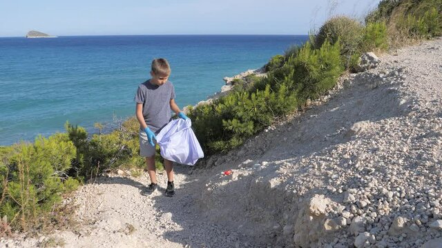 Nature cleaning, stop plastic, environmental concept. Volunteer kid raises and throws plastic garbage in the bag for recycle near the sea at sunny day
