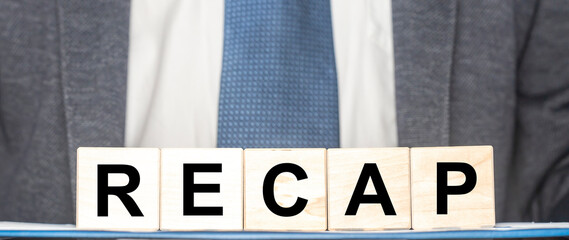 Recap word from wooden blocks on desk board with man in suit on background. Business recap concept