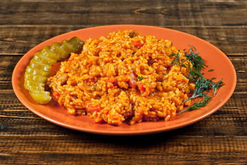 Pilaf with meat, cucumbers, greens and carrots in an orange plate. Rice on a wooden brown board.