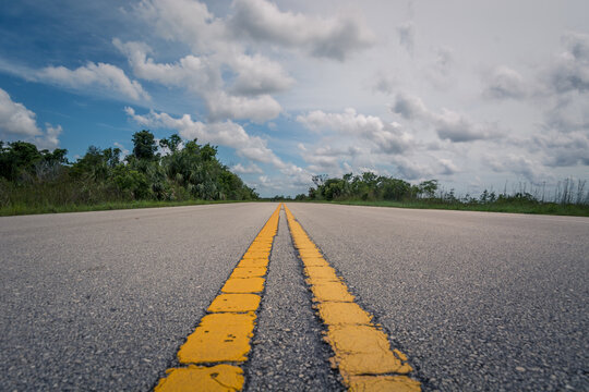 Low angle straight empty long asphalt road in Everglades National Park, Florida, USA. Two yellow lines in the middle. Blue sky with clouds. Wide shot