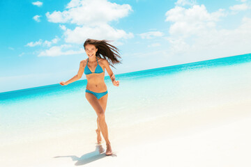 Beach fun happy Asian woman laughing running in blue bikini from ocean Caribbean travel destination. Slim body model enjoying summer holidays.