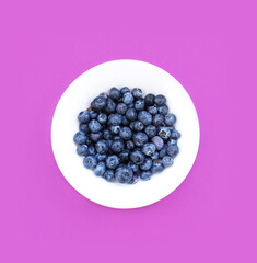 Blueberry in a white plate on a violet background. The composition of blueberry on a colored background. Healthy food. Minimalist food. Top view