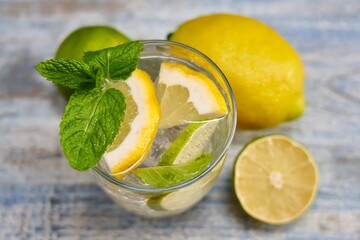 Top view image of cold refreshing summer limonade, mojito or gin tonic with fresh mint and ice cubes, lime and lemon on colorful wooden table.