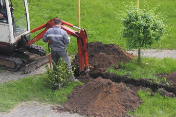 A worker in gray overalls stands next to a working compact rubber tracked excavator, which...