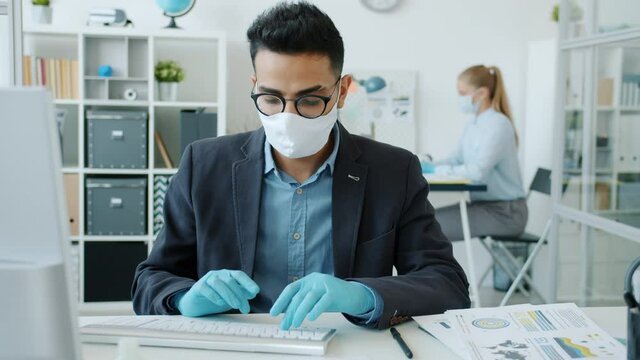 Middle Eastern man wearing mask and gloves is working with computer in office room during lockdown during corona virus global epidemic. Business and health concept.