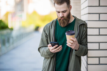 Young bearded man having walk in the city and typing sms while drink coffeea