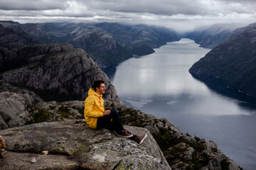 Brunet man hiker in yellow raincoat smiles and sits on the cliff of Preikestolen mountain (Preacher's Pulpit or Pulpit Rock) with background of Lysefjord and low cloudy sky in Norway