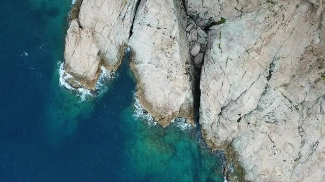 Aerial shot of waves splashing on rock formation in blue sea, drone ascending over ocean - Tossa de Mar, Spain