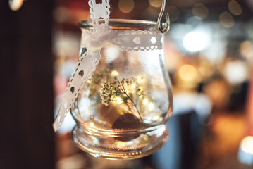 Hanging round glass container used as vase decorated with white ribbon with hearts. Abstract blurred background.