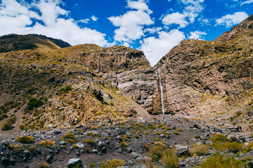 Landscape of a waterfall over Andes Mountains in a valey of Chile with blue sky, some clouds and amazing view of rock mountains great for adverture, hikking and trail run
