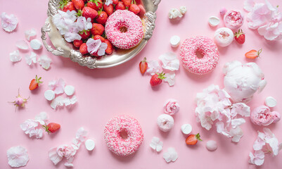 Top view composition of strawberry tray, three donuts with icing, marshmallows and pink flowers on a pastel pink background. Festive concept.