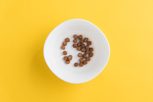 White bowl with few chocolate corn cereal balls on yellow background, top view