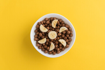 White bowl full of chocolate cereal balls and banana slices on yellow background