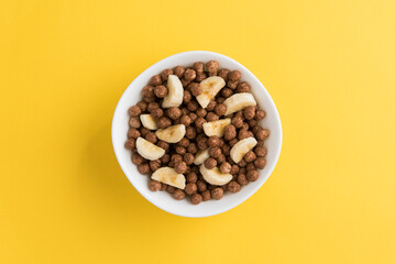 White bowl full of chocolate cereal balls and banana slices on yellow background