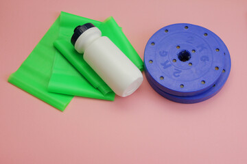 online sports at home,during self-isolation, keep the figure in shape, on a pink background is a green elastic band for exercise, a white water bottle and dumbbell discs