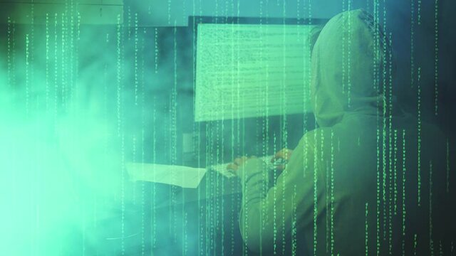 Hacker Attack. Hacker Enter to the green matrix. Hacker in a hood looking to the monitor with projected code numbers and characters. Smoke on the background