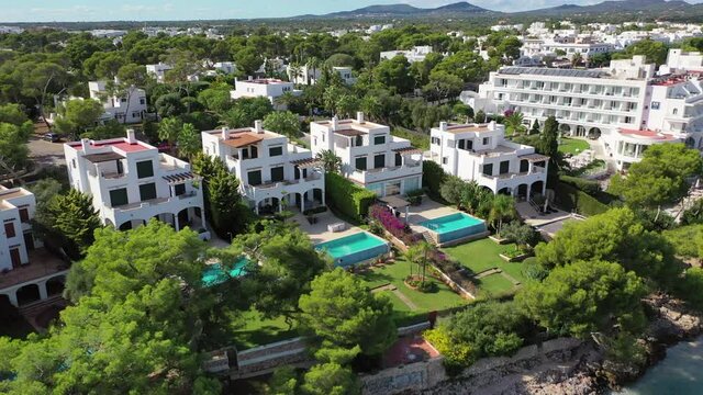 Aerial panning shot of resorts with swimming pools by sea on sunny day, drone flying over coastal city - Cales de Mallorca, Spain