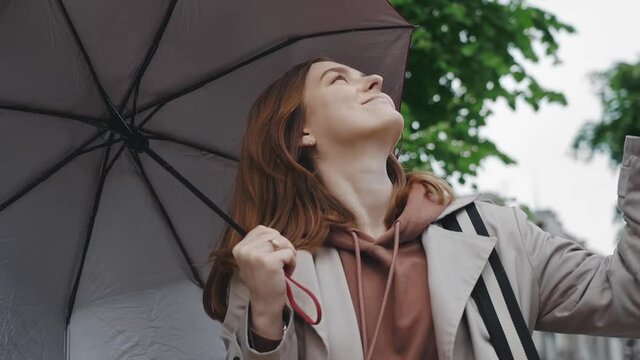 A cute young girl in a raincoat under an umbrella happily walks down the street, succumbing to rainy weather. slow motion