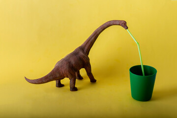 Obraz premium Dinosaurio Herbívoro bebiendo de un vaso de plástico verde con una pajita en un fondo amarillo