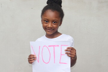 African American child holding white paper sign with word Vote in pink letters standing outdoors