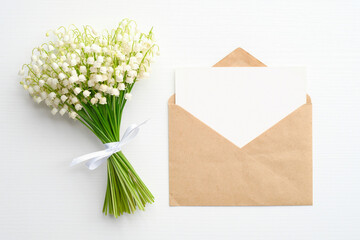 Wedding invitation or love letter concept. Bouquet of lily of the valley flowers and envelope with blank paper card mockup on white table. Flat lay, top view.