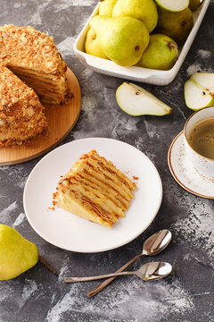 A piece  of pear layered cake on white plate with fresh pears, whole cake and coffee in background