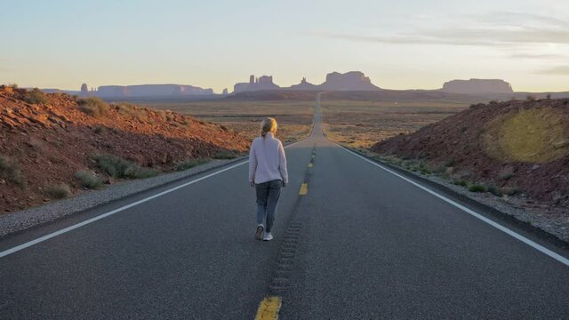 young woman in a fluffy white sweater walks along an empty deserted mountain road on a background of red mountains Monument Valley long shot