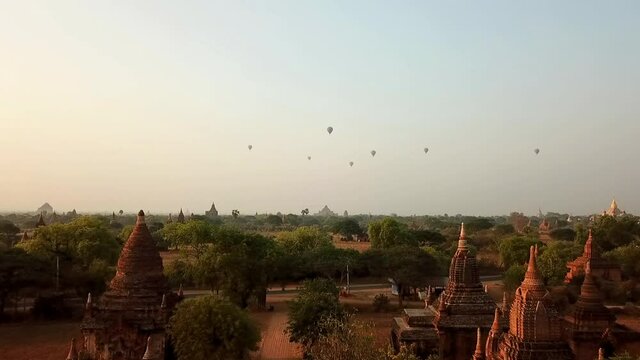 Aerial shot of hot air balloons over pagodas and field, drone ascending forward over Buddhist temples amidst trees against sky - Bagan, Myanmar