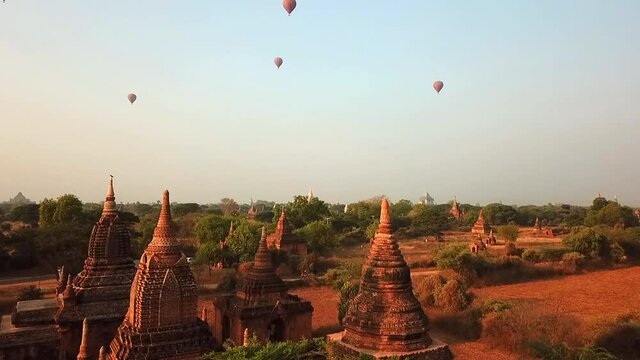 Aerial shot of hot air balloons flying over pagodas on sunny day, drone ascending over Buddhist temples amidst trees against sky - Bagan, Myanmar