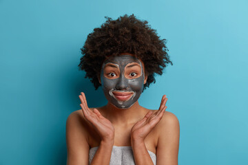 Positive happy dark skinned Afro American woman applies facial clay mask, gets beauty treatments, cares abot skin, spreads palms sideways over face, stands wrapped in towel, models indoor. Hygiene