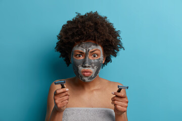 Serious woman with Afrohair concentrated at something very attentively, applies clay mask for reducing wrinkles, holds razor going to have hygienic procedures after taking shower isolated on blue wall