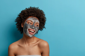 Joyful positive dark skinned curly woman stands naked indoor, applies beauty mud mask for perfect...