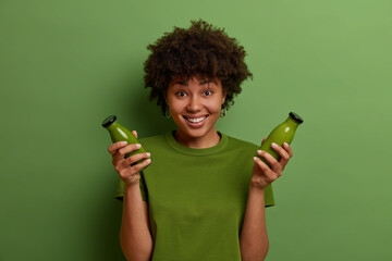 Happy dark skinned girl holds glass bottles with raw green detox vegetable smoothie, leads healthy...