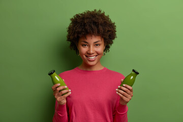 Portrait of happy smiling dark skinned woman has healthy natural lifestyle, holds two bottles of green vegetable smoothie, has proper nutrition, enjoys weight loss drink, wears rosy jumper, smiles